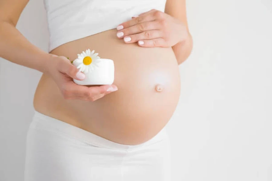 Woman's Hand Holding White Cream Jar with Chamomile on Big Naked Belly