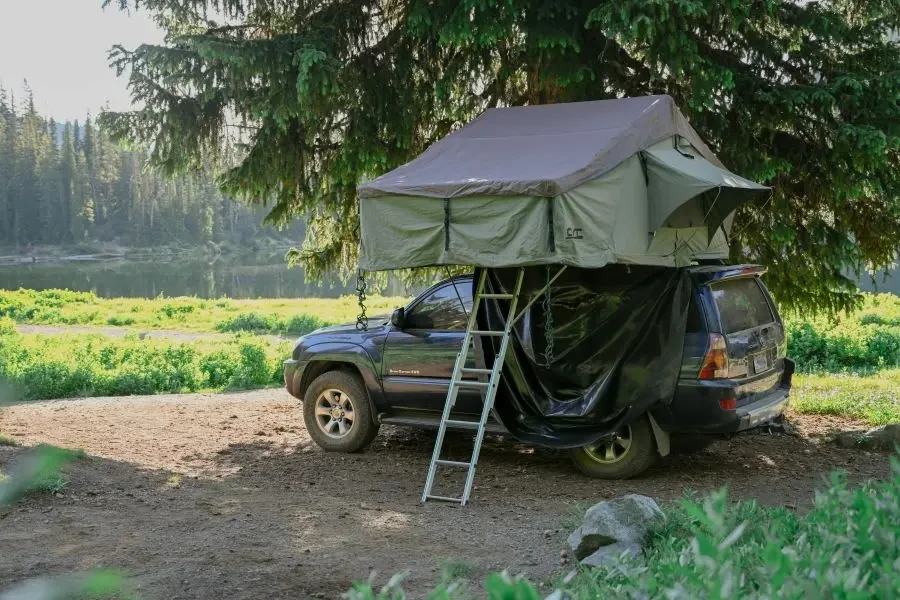 A car equipped with a rooftop tent is parked in a serene forest campsite by the lake