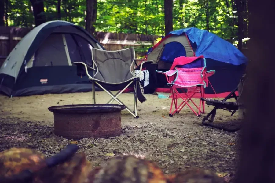 A serene campsite in the woods with colorful tents, camping chairs, and a fire pit, perfect for a leisure getaway