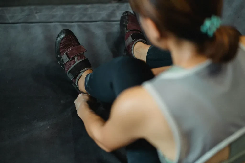 A female climber adjusts her shoes while sitting on a mat in a gym