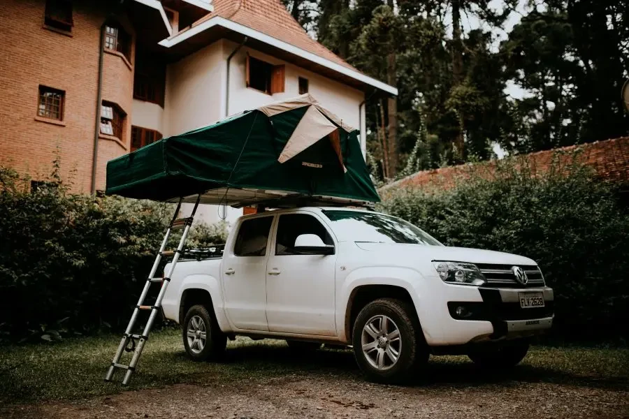 White pickup truck with roof tent and ladder, parked by a rustic brick building surrounded