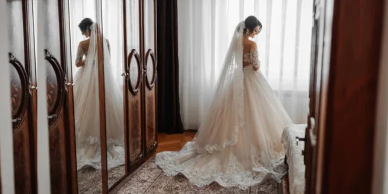 A Brunette Bride in a White Dress and a Long Veil Poses Standing by the Window