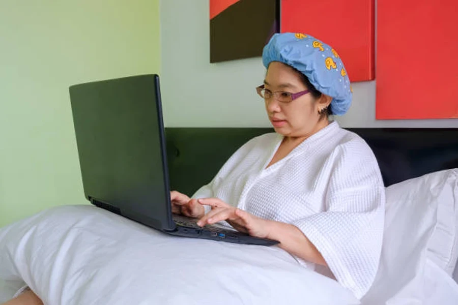 Asian Middle Aged Woman in White Bathrobe with Shower Cap Using Laptop on Her Bed