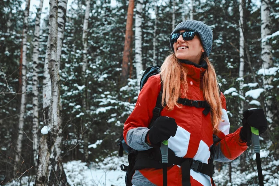 Beautiful Young Woman Dressed in Warm Sportswear, Hat and Sunglasses Stands with Trekking Poles in a Snowy Pine Forest