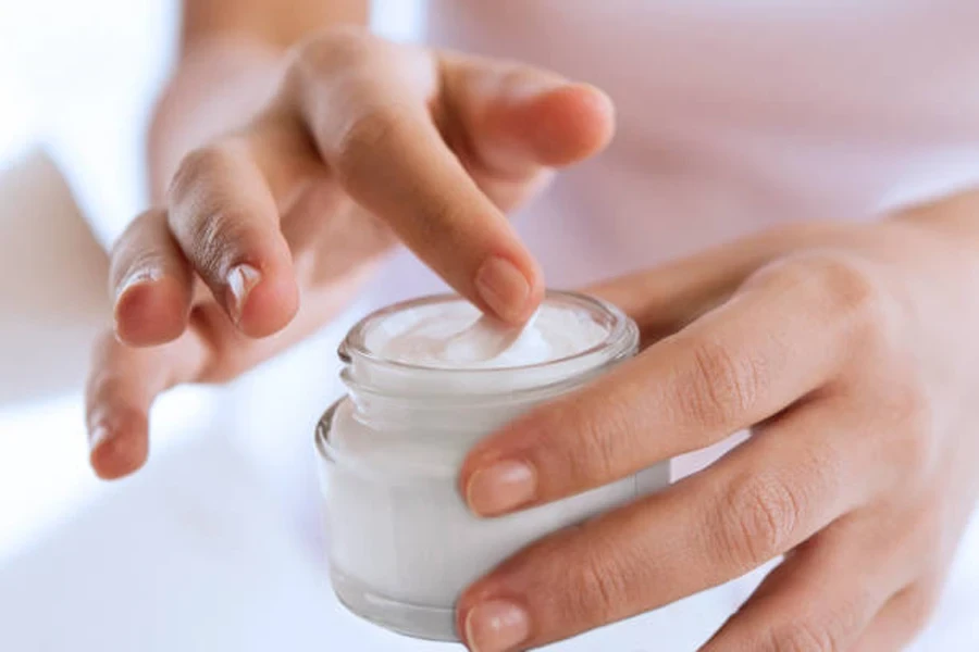 Finger scooping out white hand cream from glass jar