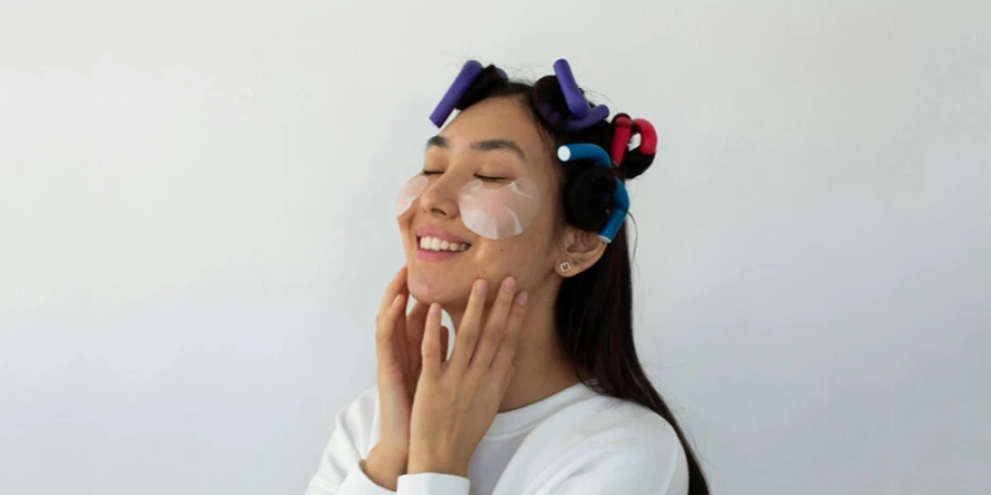 Smiling young ethnic female with hair rollers touching cheeks with eye patches on white background