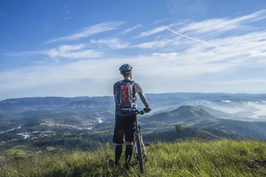 A cyclist stands on 1_a hill, gazing at a stunning mountain landscape on a bright sunny day