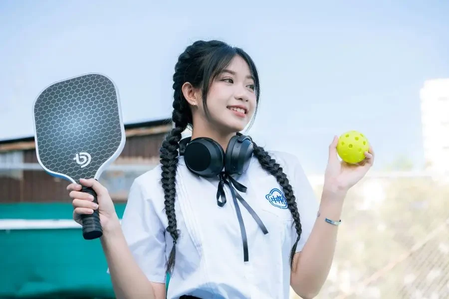 Smiling Asian woman holding a ball and paddle in outdoor setting. Urban sports vibe