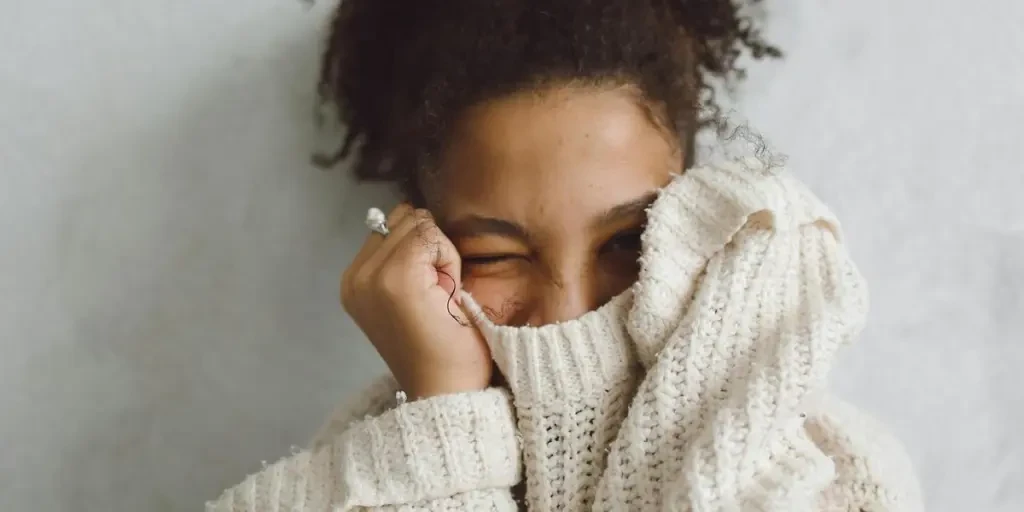 A cheerful woman winks while partially covering her face in a warm cream sweater