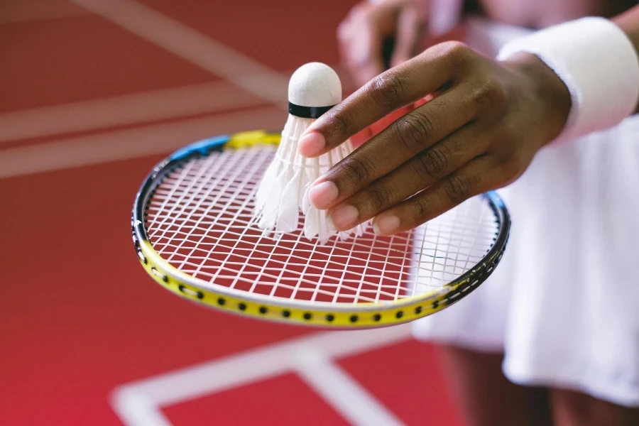 A detailed view of a hand holding a shuttlecock over a badminton racket, on an indoor court