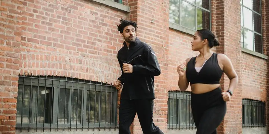 A man and woman in athletic gear jogging beside a brick wall outdoors, expressing energy and communication