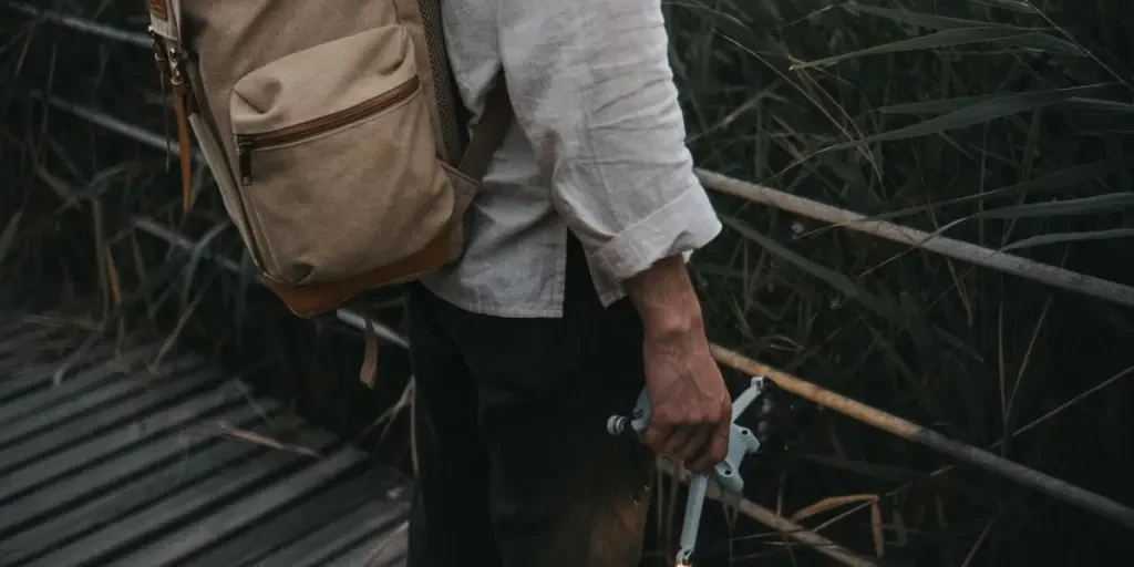 A man wearing a backpack stands on a wooden walkway holding a drone, surrounded by tall grass