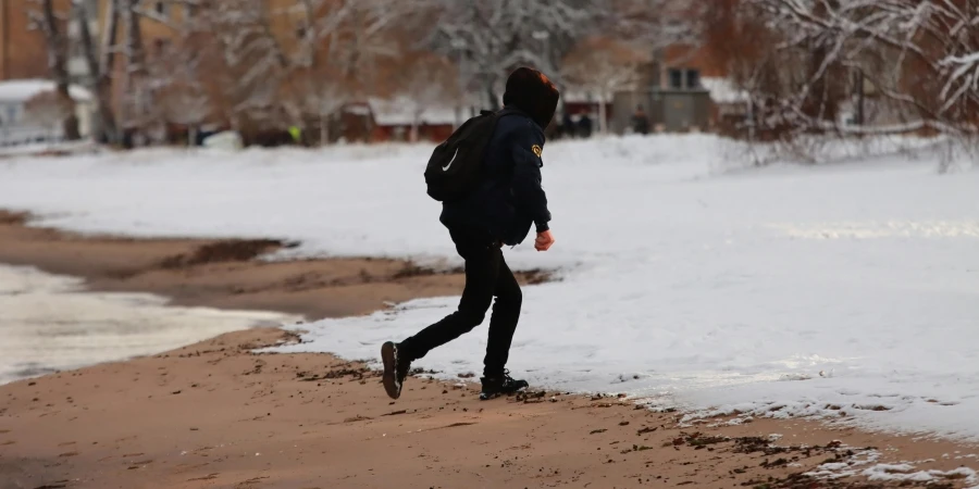 A person walks along a snowy beach during winter near residential buildings