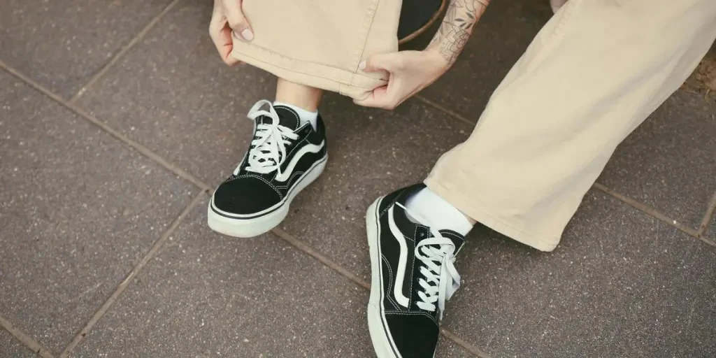 A person with a tattoo adjusts their beige pants, showcasing black sneakers on a paved surface