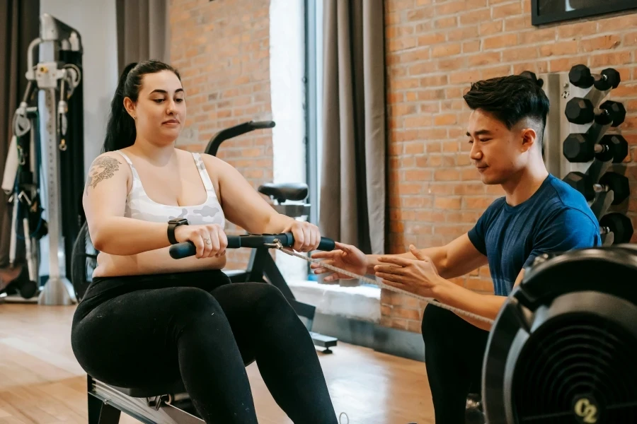 A personal trainer assists a woman on a rowing machine in a gym environment