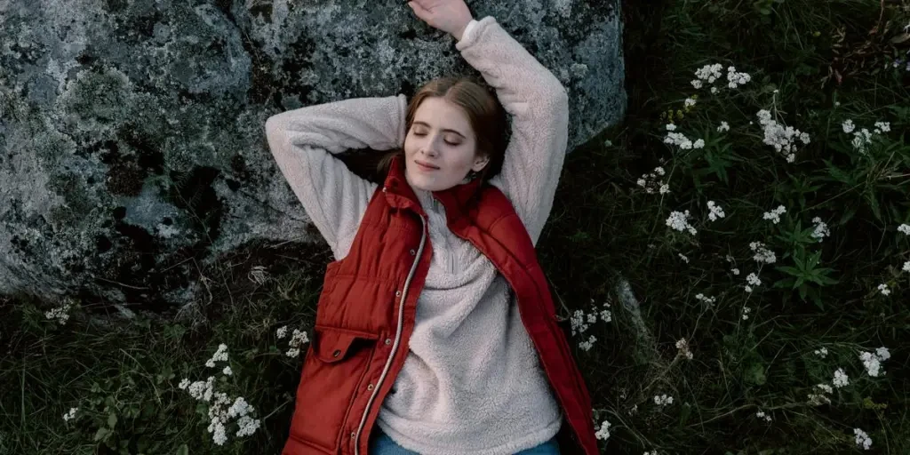 A serene scene of a young woman lying outdoors on grass, eyes closed in relaxation, surrounded by flowers