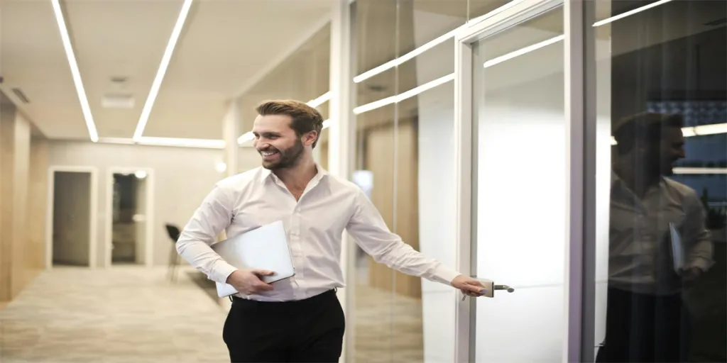 A smiling man holds a laptop while standing in a modern office hallwayexuding confidence by Andrea Piacquadio