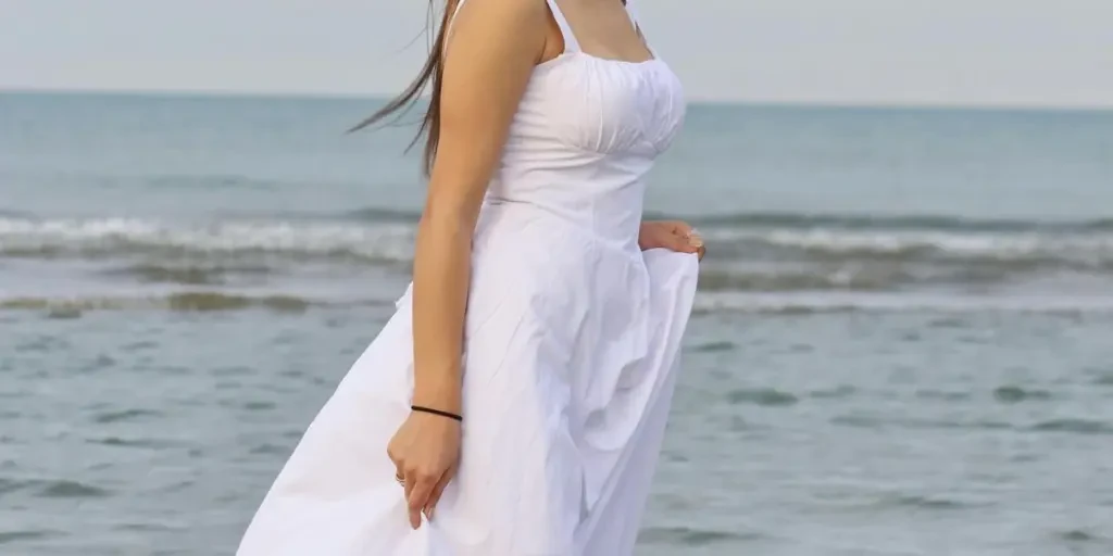 A woman in a long white dress standing ankle-deep in the ocean during summer