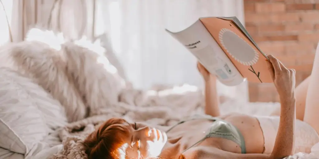 A woman in lingerie reading a magazine while lounging on a bed, enveloped in soft, natural light