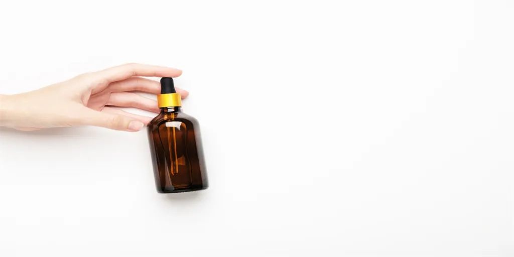 A woman's hand holds a glass brown bottle with pipette with essential oil on white background