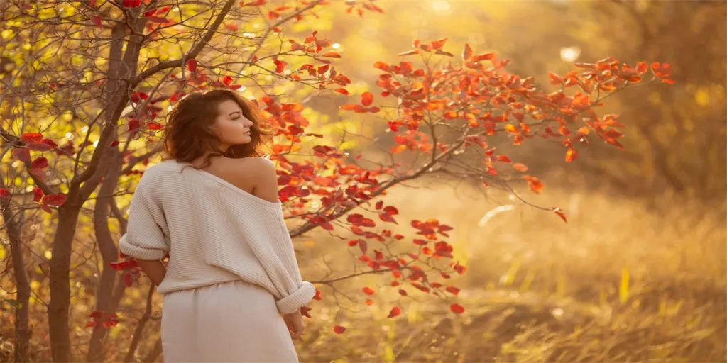 A young brunette woman in a cozy off-shoulder sweater poses near a tree with vibrant autumn leave