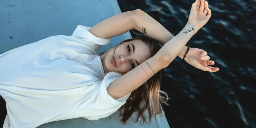 A young woman in a white t-shirt poses artistically by the ocean, exuding a sense of relaxation