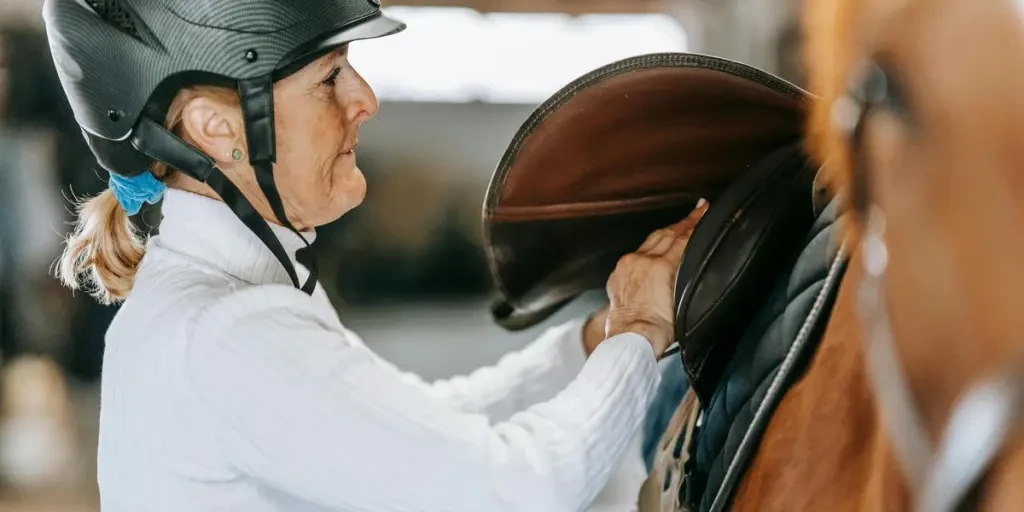 Adult woman prepares horse for riding by adjusting saddle in stable