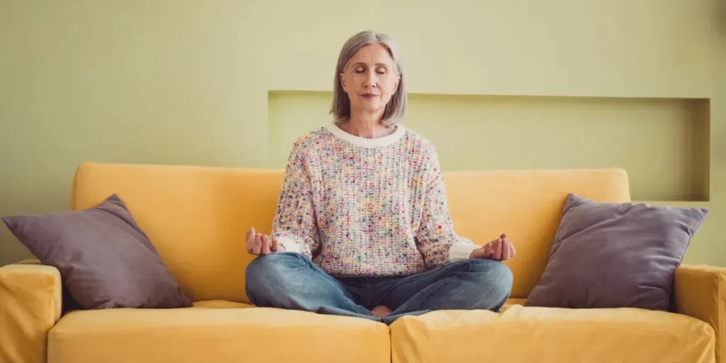 An elderly woman meditates peacefully on a yellow sofa