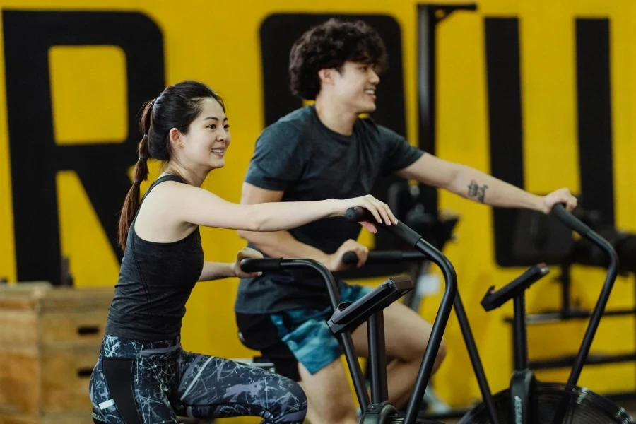 Asian man and woman enjoying a workout on stationary bikes in a gym, smiling and energetic