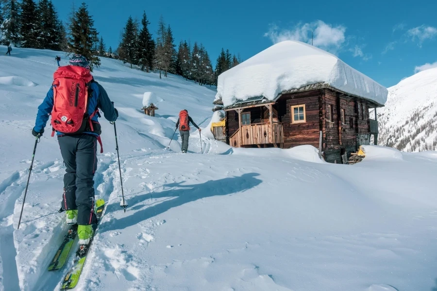 Backcountry skiers approach a rustic snow-covered cabin in a picturesque alpine setting