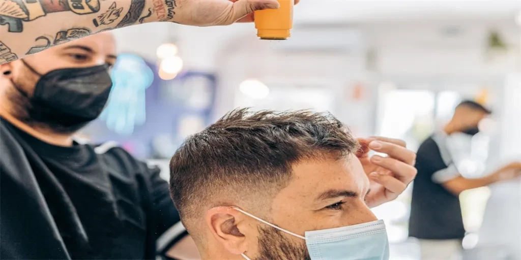 Barber throwing talcum powder on the hair of a caucasian man wearing a mask in a barbershop