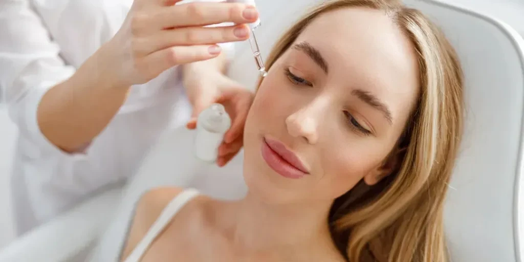 Close up of beautician hand holding bottle of essential oil and pipette while pouring skincare serum on woman cheek in beauty salon