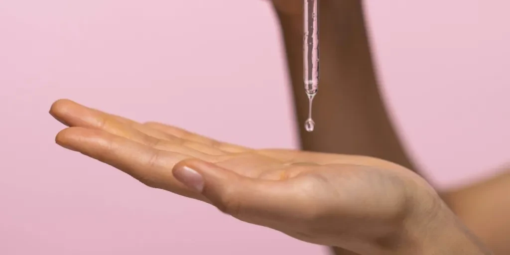 Close-up of hands with dropper applying face serum against a pink background for skincare routine