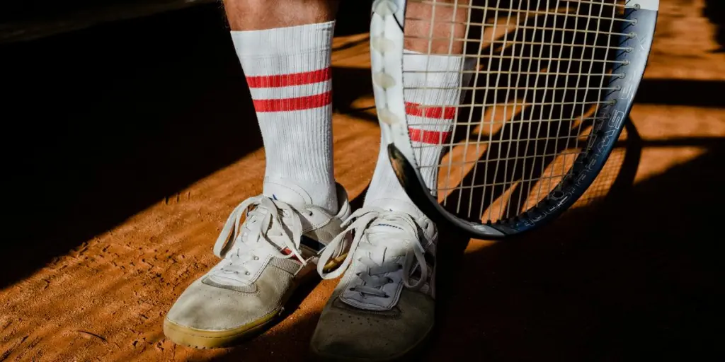 Close-up of tennis shoes, socks, and a racket on a clay court with shadows