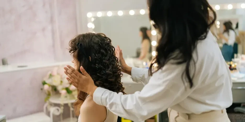 Close-up side view of hairdresser applying hair styling product to female client curly hairstyle in beauty studio