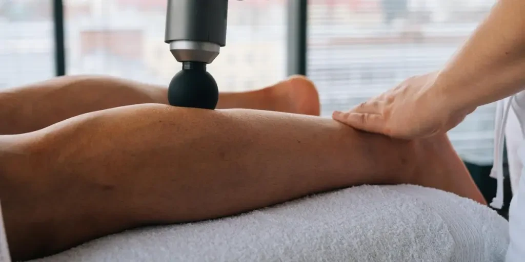 Close-up side view of unrecognizable professional male masseur massaging leg calf muscles using massage gun percussion tool of muscular athlete man