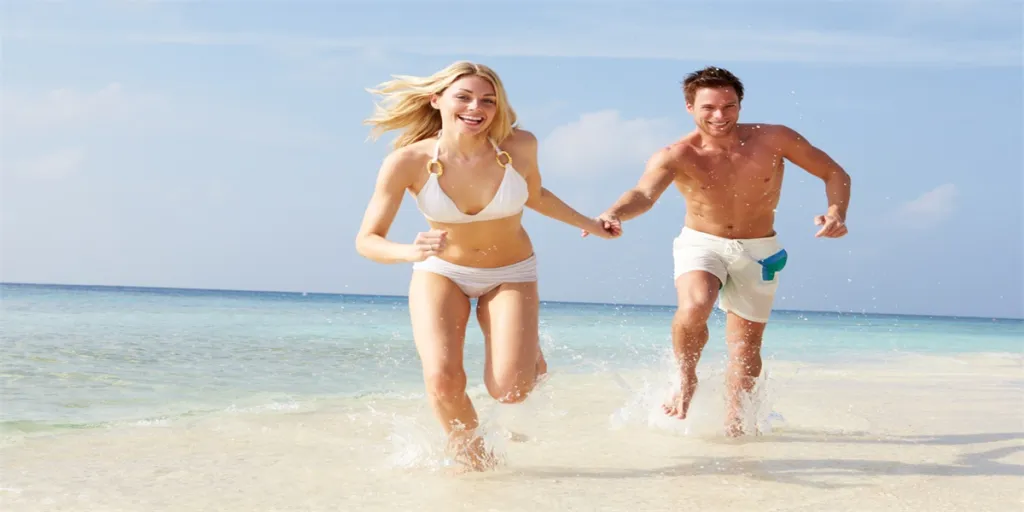 Couple Running Through Waves Towards Camera On Beach Holiday