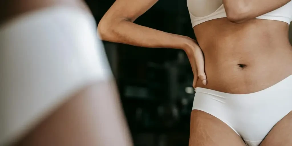 Crop unrecognizable slim ethnic female in bikini and bra standing with hand on waist against mirror