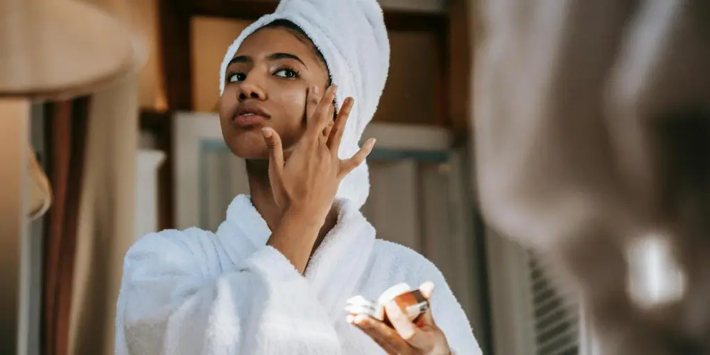 Crop young ethnic female in bathrobe with terry towel on head applying smooth cream on face while looking in mirror at home