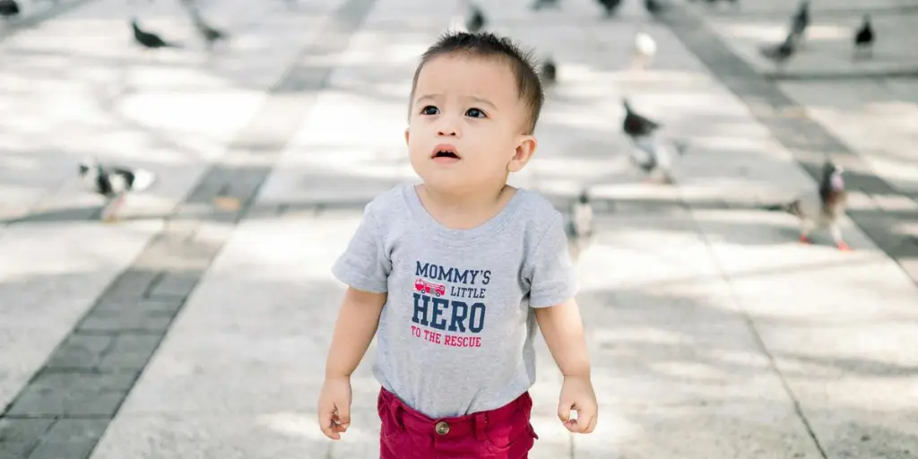 Cute baby boy in a park surrounded by pigeons, wearing a t-shirt with text, under a sunny sky