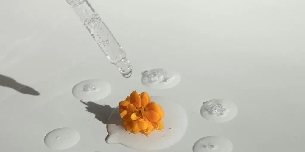 From above of drops of transparent moisturizing cosmetic product dripped by pipette and small fresh flower head placed on white table