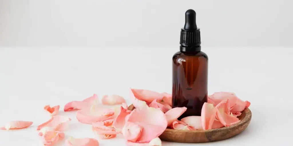 Gentle pale pink wavy rose petals placed on small round wooden plate and table near dark glass essence flask on white background by Photo