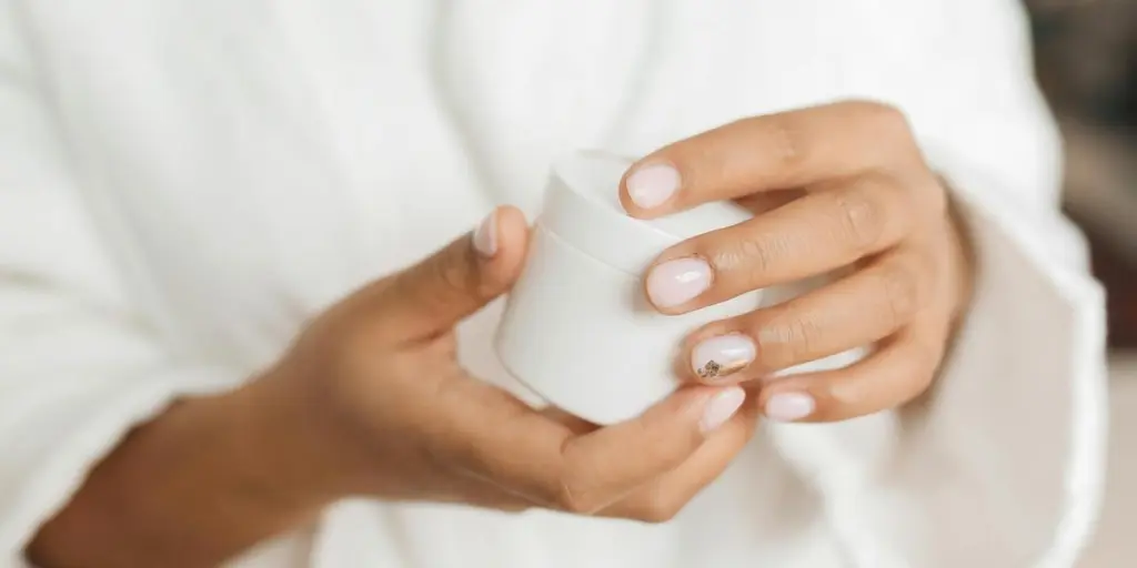 Hands with manicured nails holding a skincare container in a bathrobe, close-up view