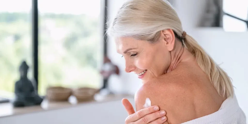 Mature woman in white bathrobe applying moisturizer cream on shoulder and body after shower