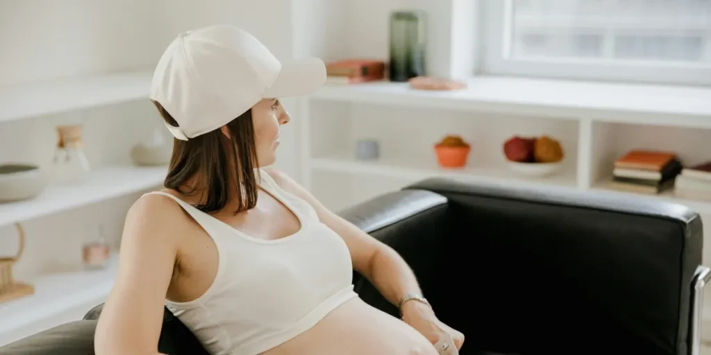 Pregnant woman in a white cap rests on a black sofa, showcasing her baby bump in a bright indoor setting