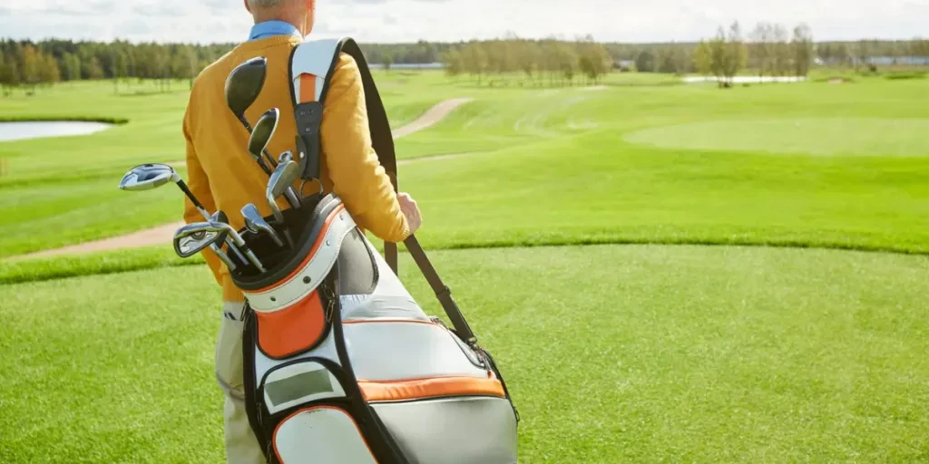 Rear view of mature golfer with sportive bag and bunch of clubs standing in front of vast green play field