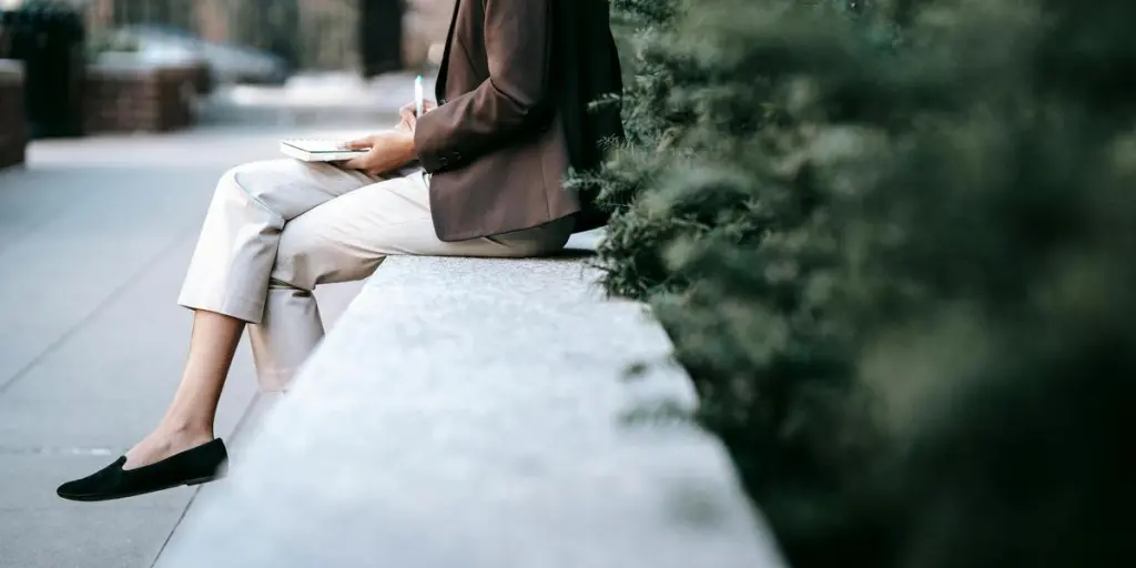 Side view of crop unrecognizable young ethnic female employee in classy outfit sitting on stone border in city park and taking notes in planner