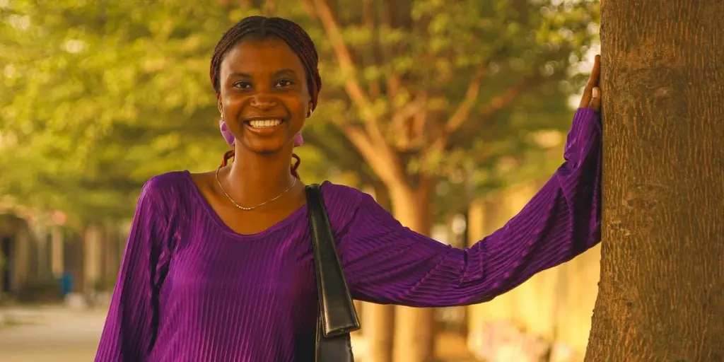 Smiling woman wearing a purple dress leans on a tree in a serene outdoor setting