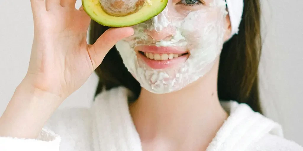 Smiling woman with face mask holding avocado for skincare in spa setting