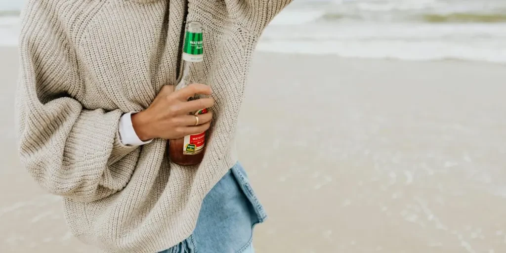 Smiling young woman in a cozy sweater holds a beer bottle at the beach, enjoying a relaxing day by Photo